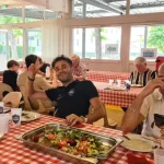 Adhésion au club l'échiquier Agenais, un moment de détente improvisé dans un espace repas pour joueur d'échecs. Les tables sont recouvertes avec une nappe à carreaux blanc et rouge. Les convivent mangent et il ont à leur disposition de grand plats de b nourriture et des gobelets à l'effigie du club. Au premier plan un monsieur portant une chemisette blanche consulte son téléphone, et un autre monsieur portant une chemisette foncée fait un signe de sympathie en levant le bras et dressant deux doigts.