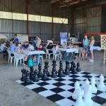 Jeu d'échec géant en premier plan avec quatre tables derrière où des enfants sont assis, trois adultes sont debout à côté des tables et trois adultes sont à droite, ils regardent l'échiquier géant