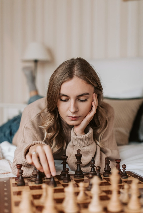 Une jeune fille aux cheveux longs châtain, allongées sur un lit avec un plateau de jeu d'échec devant elle. Elle tient dans sa main un pion noir.