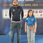 Sur un podium bleu, un homme d'une trentaine d'années aux cheveux cours, avec une paire de lunette et une barbe, portant une veste avec le logo de la FFE, se tiens debout avec à sa droite une fillette d'environ huit ans, long cheveux bruns et portant une chemisette bleu claire avec le logo "l'échiquier Agenais". Le fond de la scène est une grande affiche du championnat de France jeunes 2023.