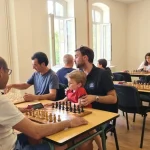 Trois tables sont installés à côté de grandes fenêtres, en premier plan quatre hommes sont installés, ils jouent aux échecs. Un homme a un enfant sur ses genoux. six autres joueurs sont répartis sur les tables