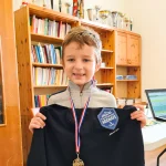 Un petit garçon avec une médaille autour du cou tient devant lui le pull siglé de l'échiquier agenais. Il y a une bibliothèque en arrière-plan