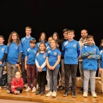 Dix huit jeunes et un adulte sont debout sur une scène sur deux rangs, ils portent tous un t-shirt bleu clair siglé du club de l'échiquier agenais