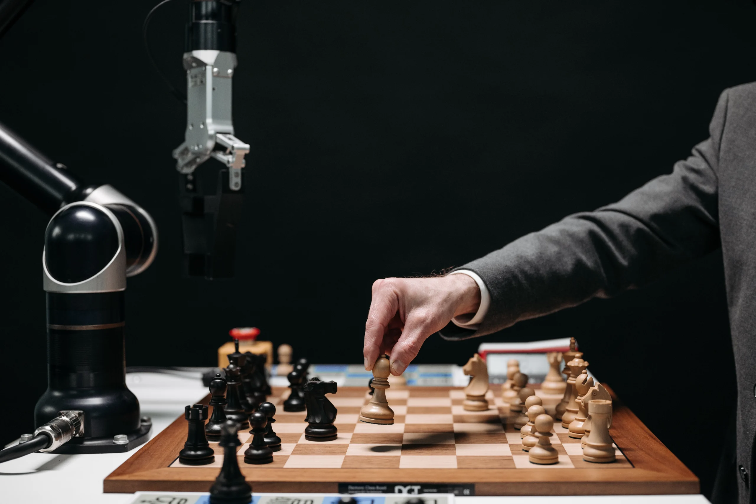 Sur la page accueil du site l'échiquier Agenais, au centre de l'image se trouve un plateau d'échecs avec à gauche une main robotisée et à droite le bras d'un homme qui saisit un pion blanc pour jouer.