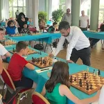 Des tables sont organisées en U, sur l'extérieur des adultes et des enfants sont assis avec chacun un plateau de jeu d'échecs devant eux. Devant un jeune garçon, un adulte de l'autre côté de la table, il est penché sur le plateau de jeu de l'enfant.