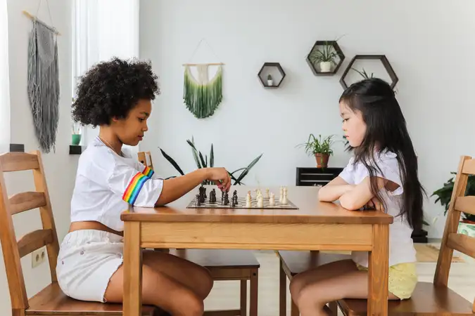 2 enfants joue aux échecs sur un table en bois. Ce sont 2 filles habillée de blanc et chacune sur une chaise en bois. Celle située à gauche à les pions noir et elle est en train de déplacer une pièce. Celle de droite la regarde les bras croisés.